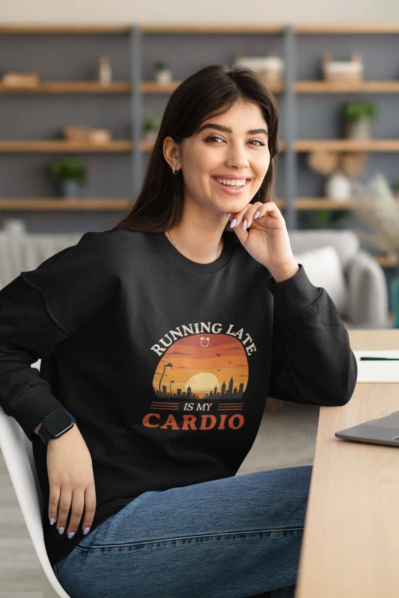 burnout humor sweatshirt with a woman wearing a black sweatshirt with a graphic design and text 'Running Late Is My Cardio', sitting in an office setting.