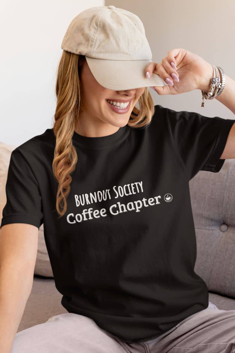 burnout humor tee with a woman wearing a black t-shirt with 'Burnout Society Coffee Chapter' text, sitting on a couch.