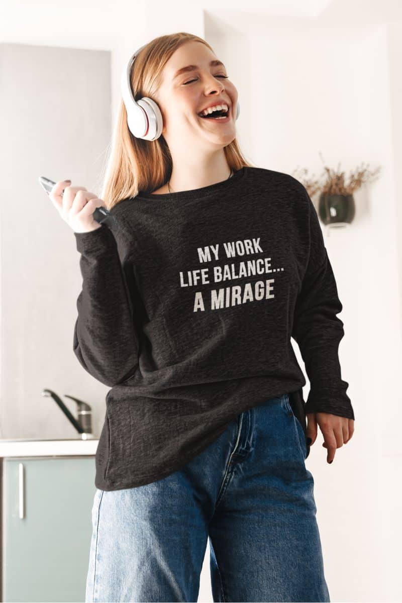 burnout recovery apparel with a person wearing a black sweatshirt with text 'My Work-Life Balance Is a Mirage', holding headphones and a phone in a kitchen.