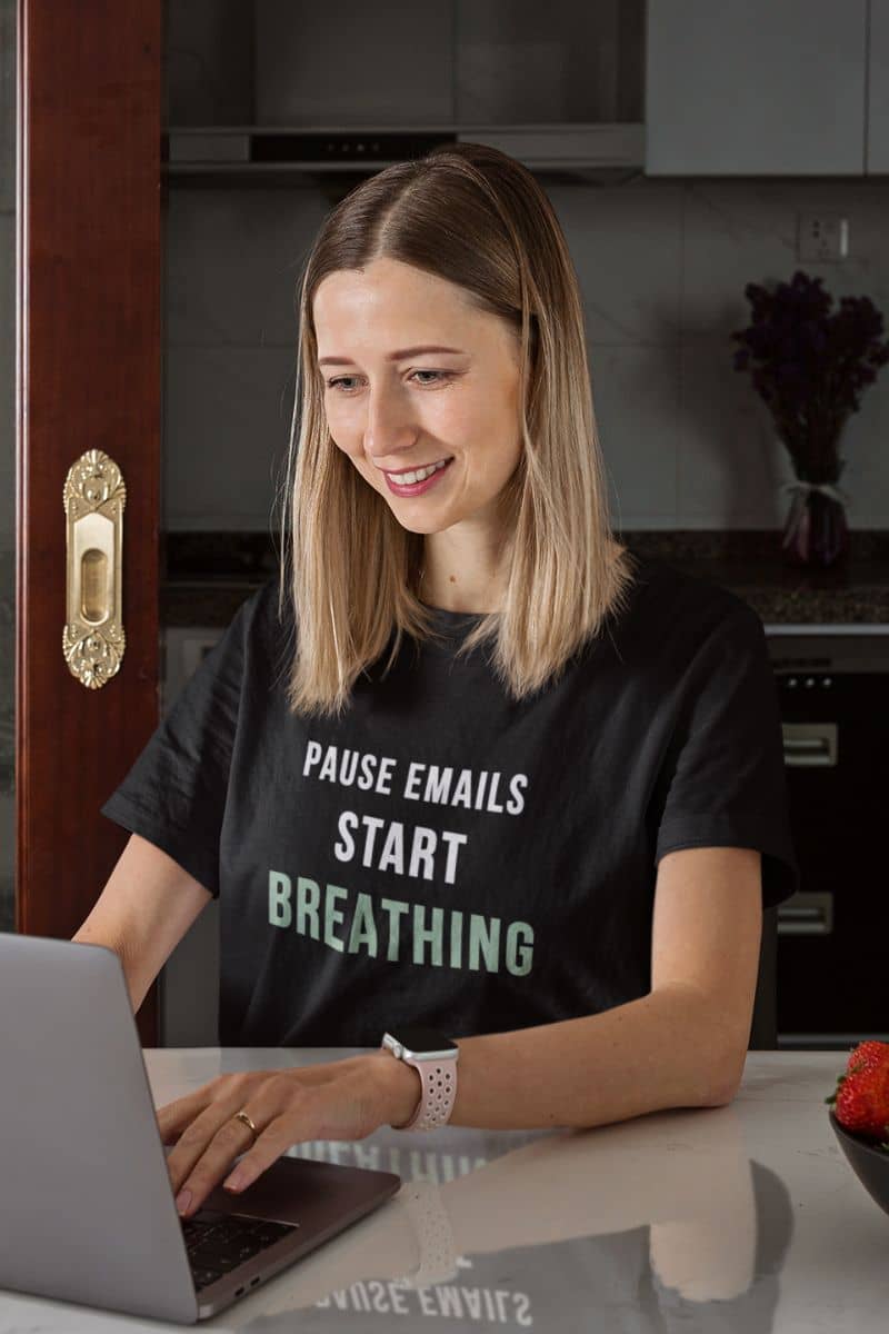 burnout recovery shirt with a woman wearing a black t-shirt with text 'Pause Emails, Start Breathing', sitting at a table with a laptop in a kitchen.
