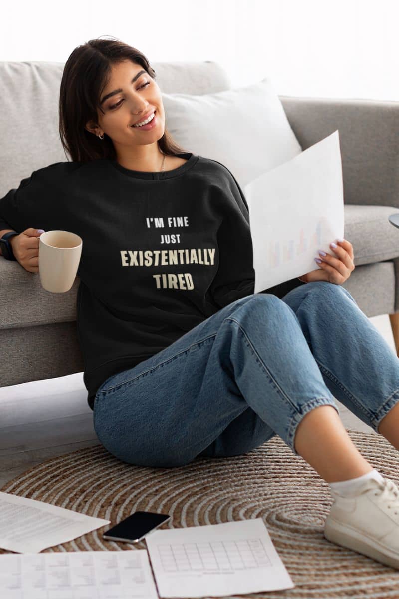 burnout recovery sweatshirt with a woman sitting on a couch wearing a black sweatshirt with text 'I’m fine. Just existentially tired.', holding a mug and a piece of paper.