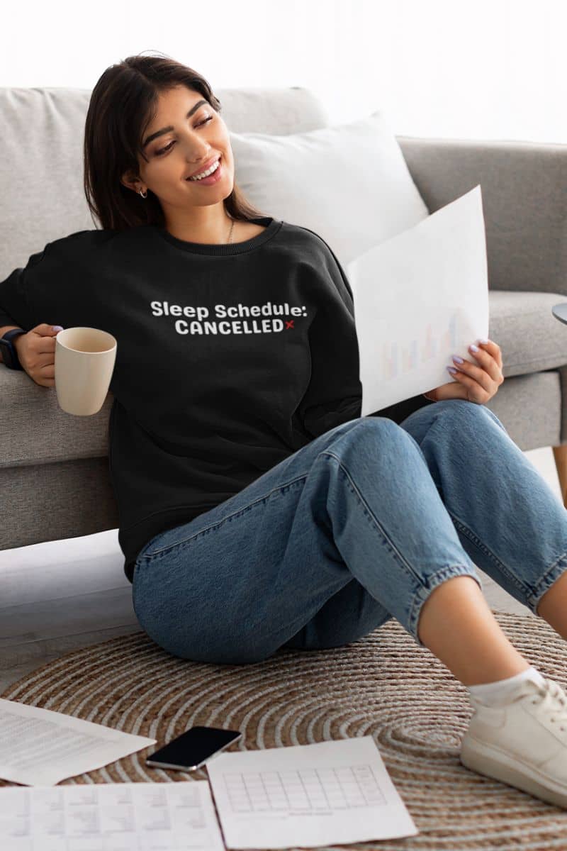 burnout sweatshirt with a woman sitting on a couch wearing a black sweatshirt with 'Sleep Schedule: CANCELLED' text, holding a coffee cup and reading a piece of paper.