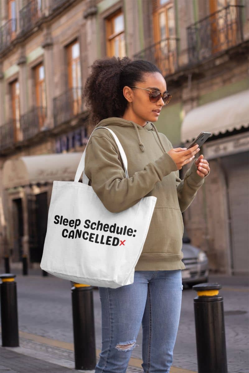 burnout tote bag with a woman holding a phone and a tote bag with text 'Sleep Schedule: Cancelled' in an urban setting