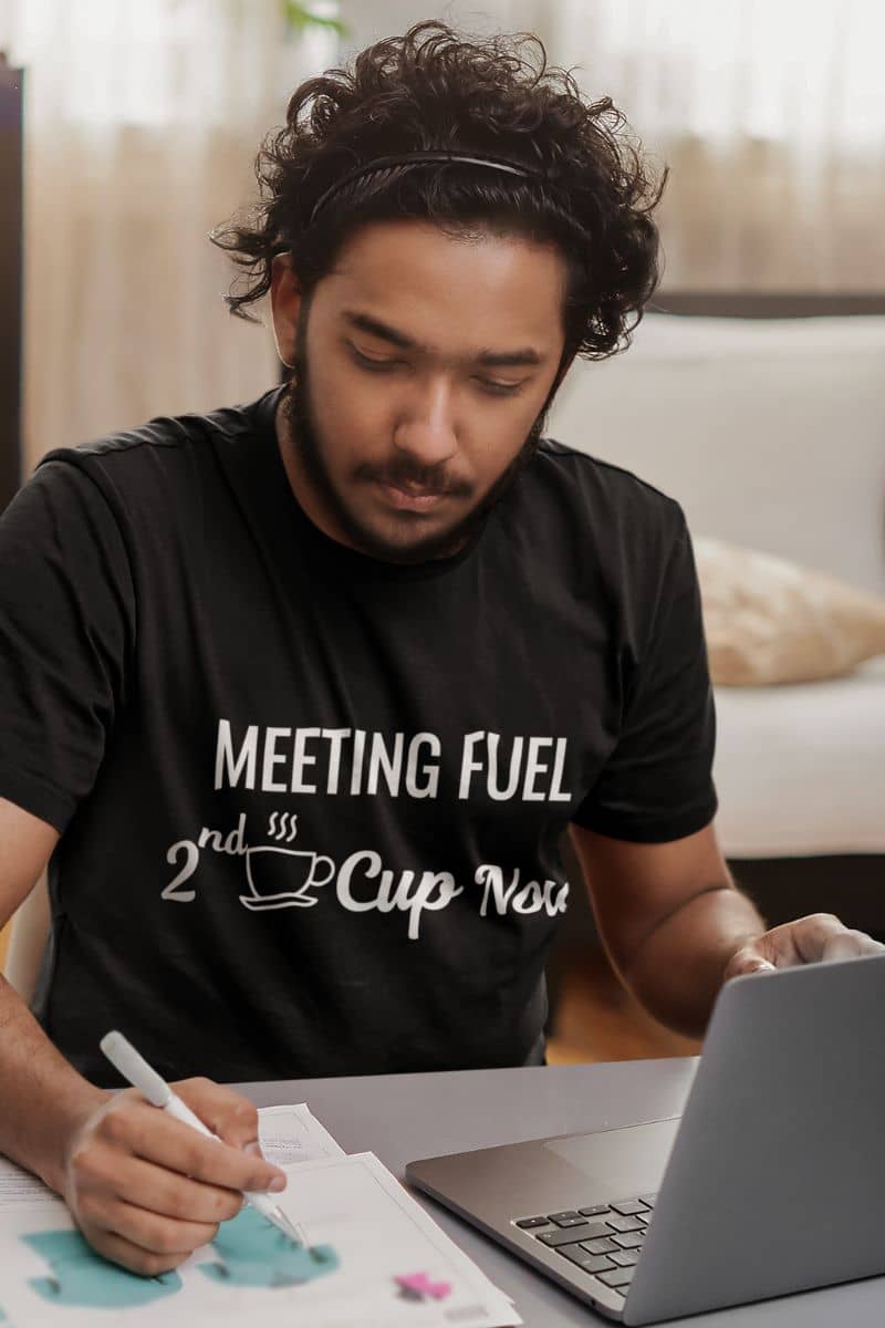 caffeine shirt with a man wearing a black t-shirt with text 'Meeting Fuel: 2nd Cup Now', sitting at a table with a laptop and paper.