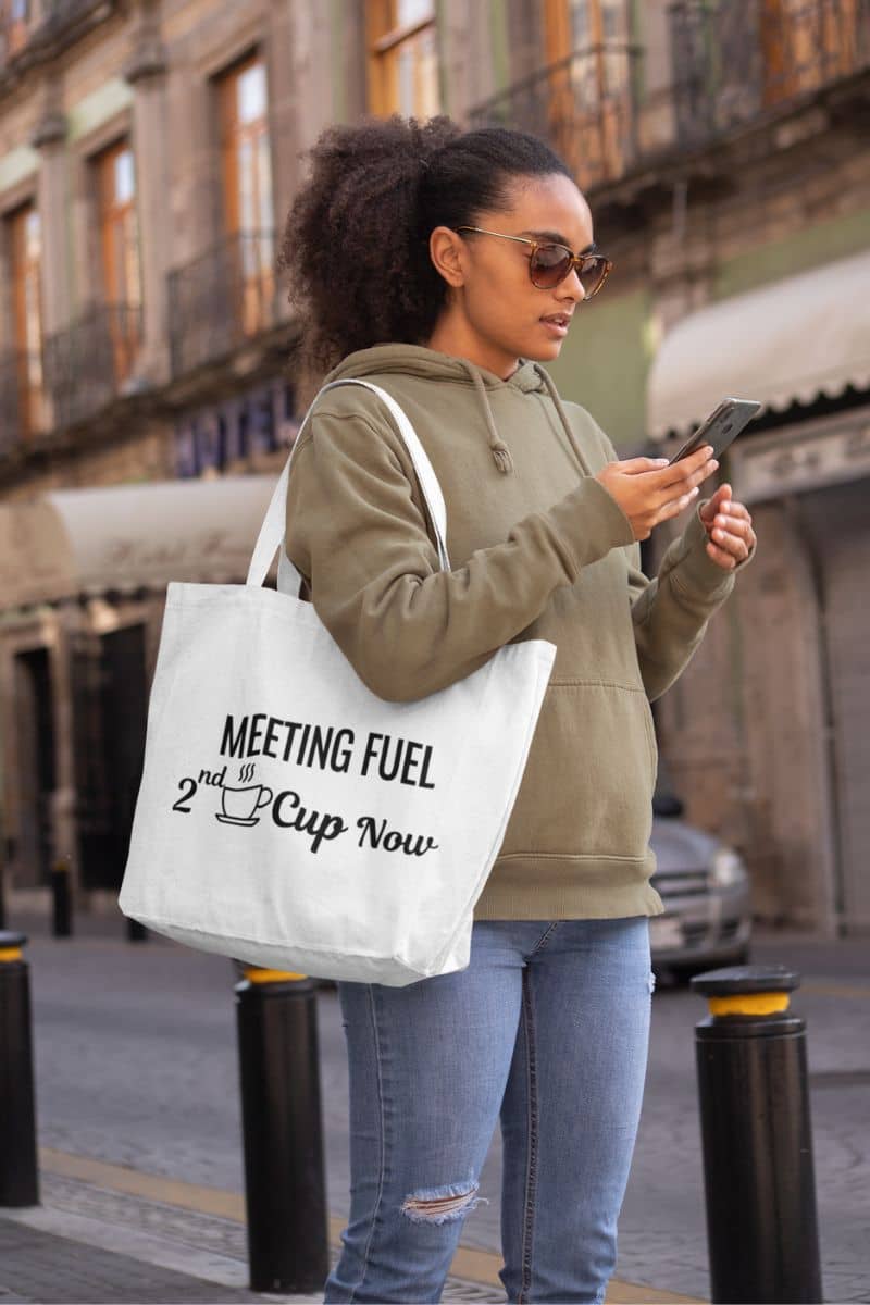 caffeine tote bag with a woman holding a tote bag with text 'Meeting Fuel: 2nd Cup Now' on a city street
