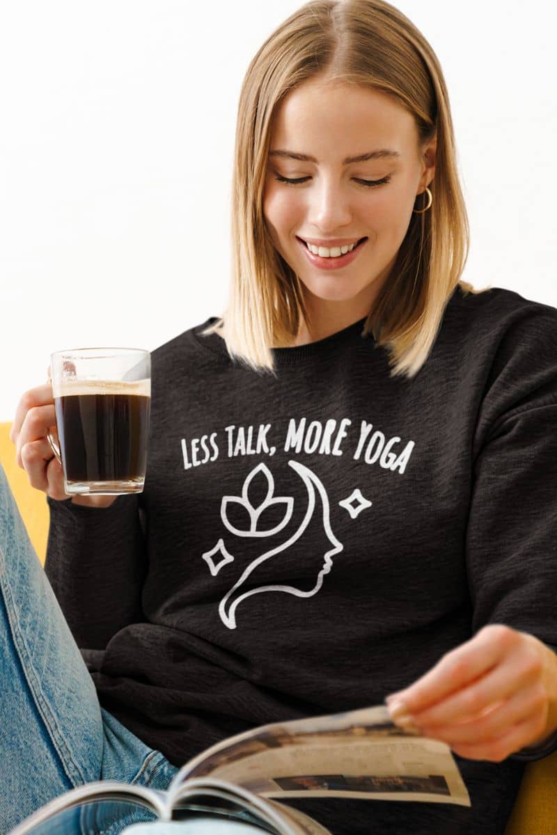 calm mindset apparel with a woman wearing a black sweatshirt with 'Less Talk, More Yoga' text and a white design, holding a coffee cup and reading a newspaper.