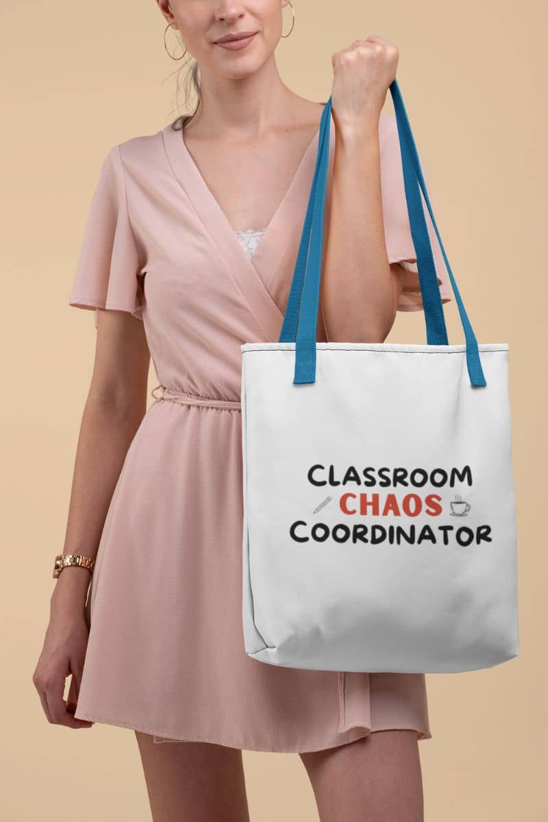 canvas teacher bag with a woman wearing a light pink dress holds a white tote bag with blue handles. The tote bag has the text 'Classroom Chaos Coordinator' printed on it in black and red lettering.