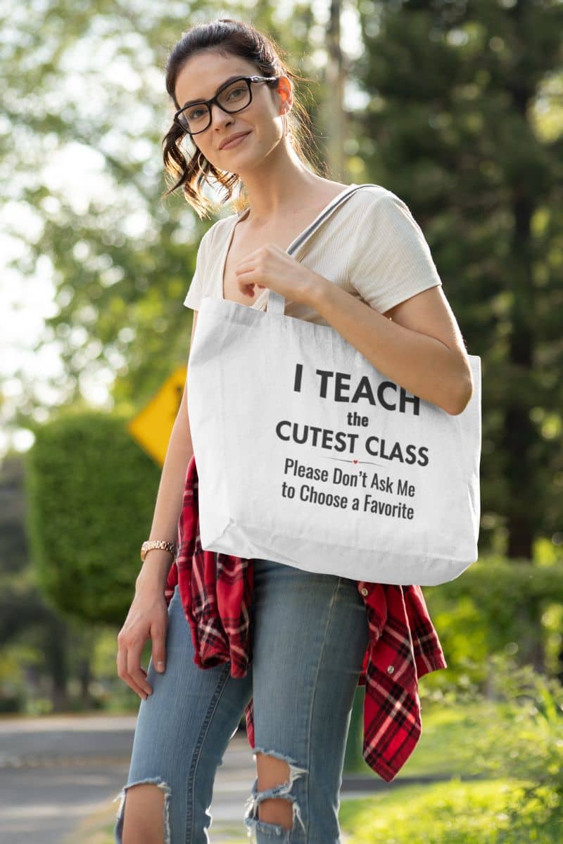 Canvas teacher bag carried by a woman outdoors, featuring black text that reads “I Teach the Cutest Class. Please Don’t Ask Me to Choose a Favorite.” She is wearing glasses, a white T-shirt, ripped blue jeans, and a red plaid shirt tied around her waist.