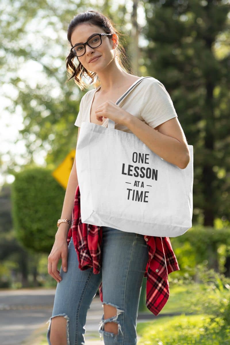 canvas teacher bag with a woman carrying a white canvas tote bag featuring the text 'ONE LESSON AT A TIME' printed in black.