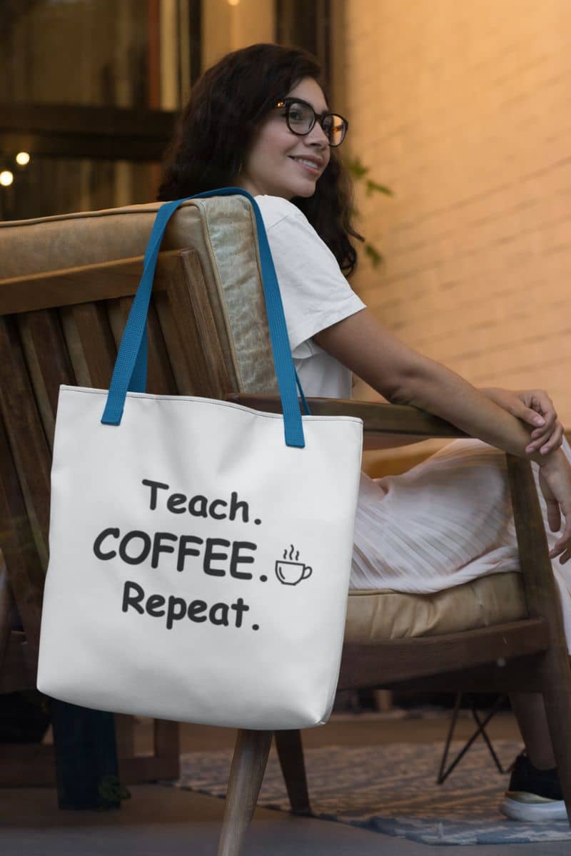 A canvas teacher tote hanging on the chair featuring the text 'Teach. Coffee. Repeat.' next to a woman sitting in the coffee shop