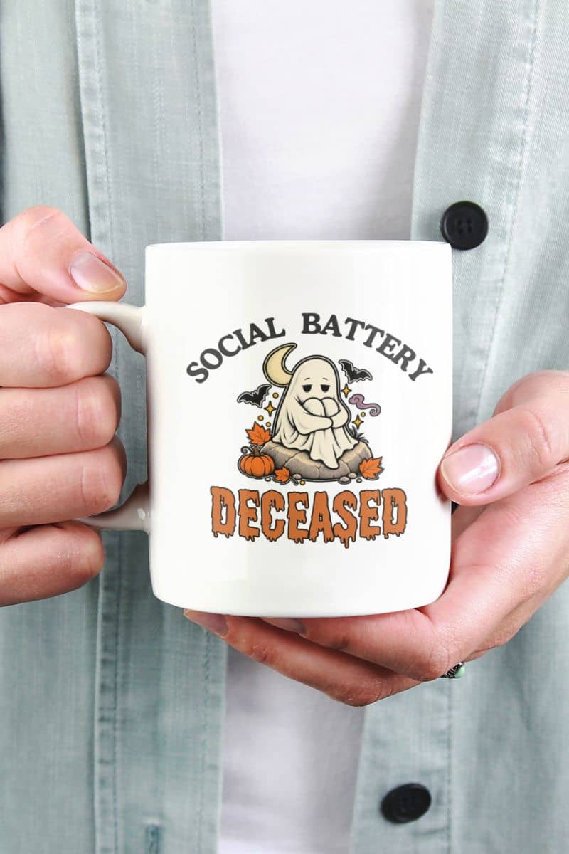 A person holding a ceramic Halloween mug with a Halloween graphic that reads 'Social Battery: Deceased' with a ghost, pumpkins, and bats.