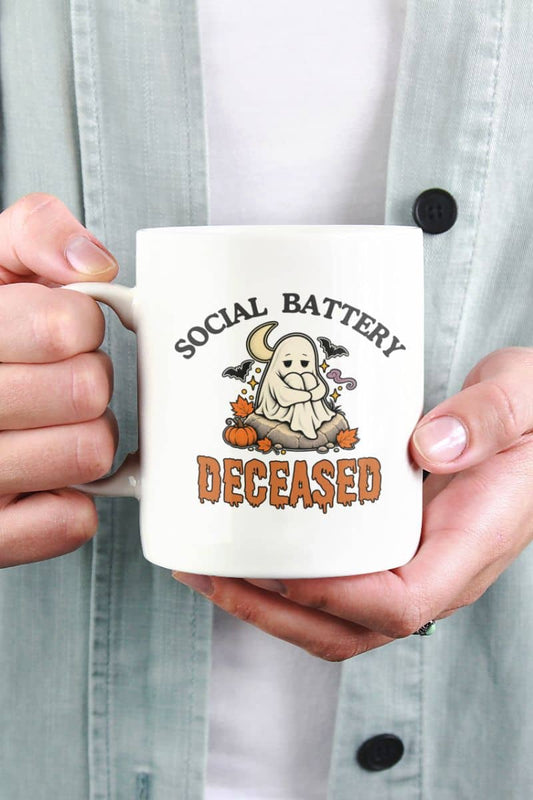 A person holding a ceramic Halloween mug with a Halloween graphic that reads 'Social Battery: Deceased' with a ghost, pumpkins, and bats.