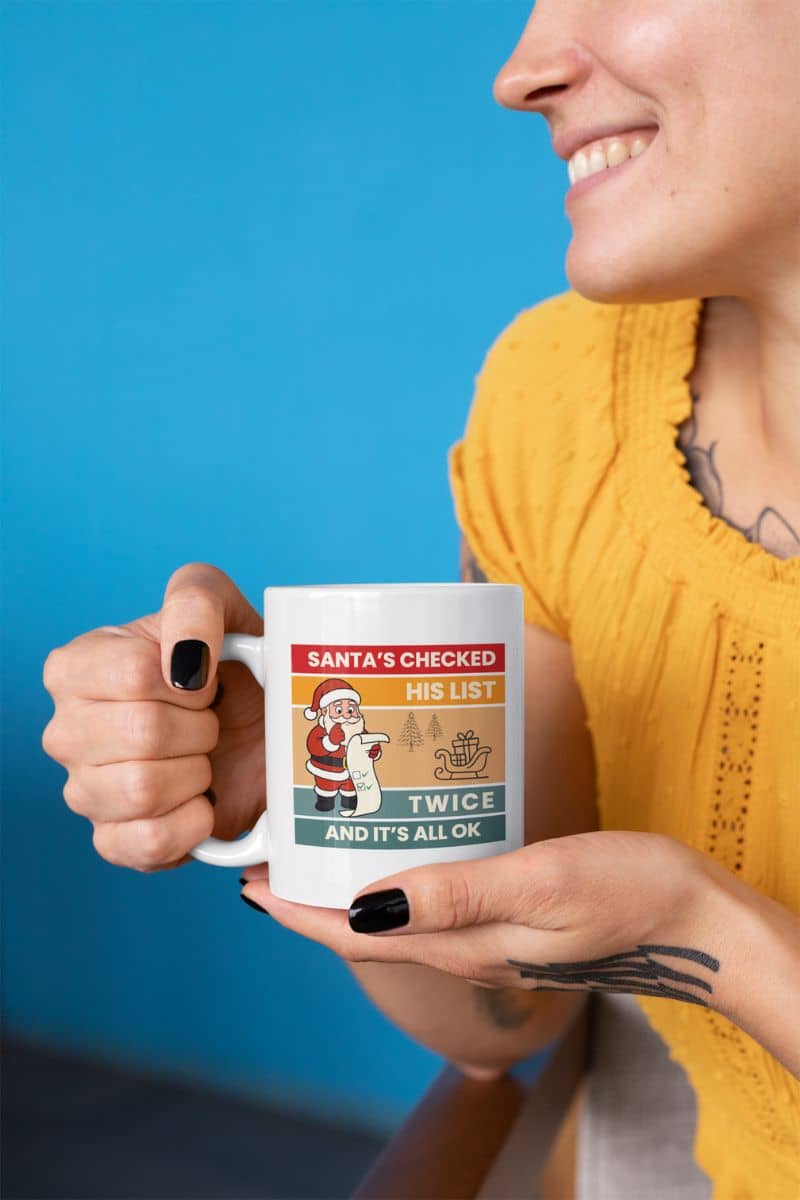 christmas coffee mug with a person holding a mug with text 'Santa's Checked His List Twice And It's All Ok' against a blue background