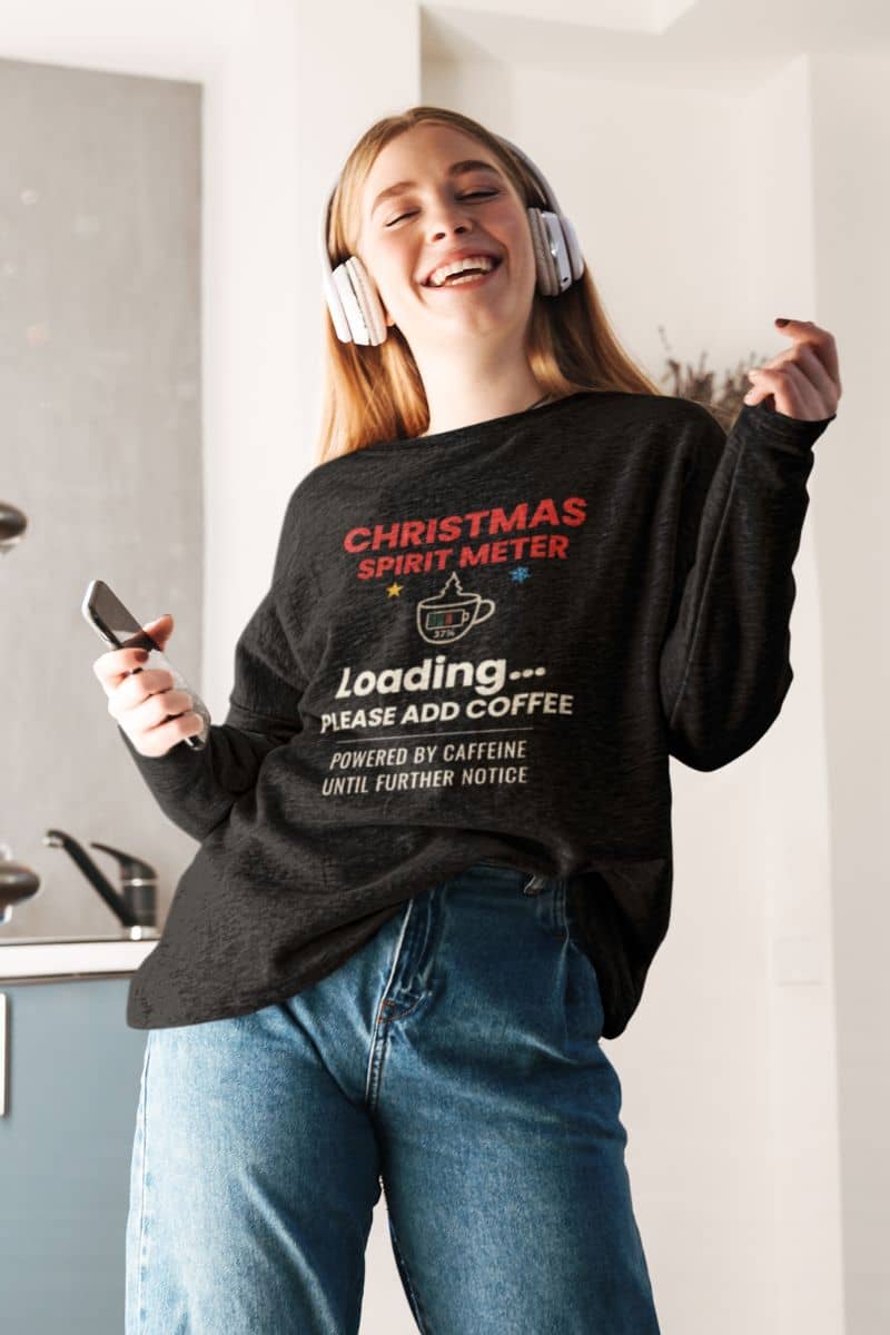 christmas coffee sweatshirt with a person wearing a black sweatshirt featuring the text 'Christmas Spirit Meter, Loading…37%. Please add coffee. Powered by caffeine until further notice' standing in a kitchen.