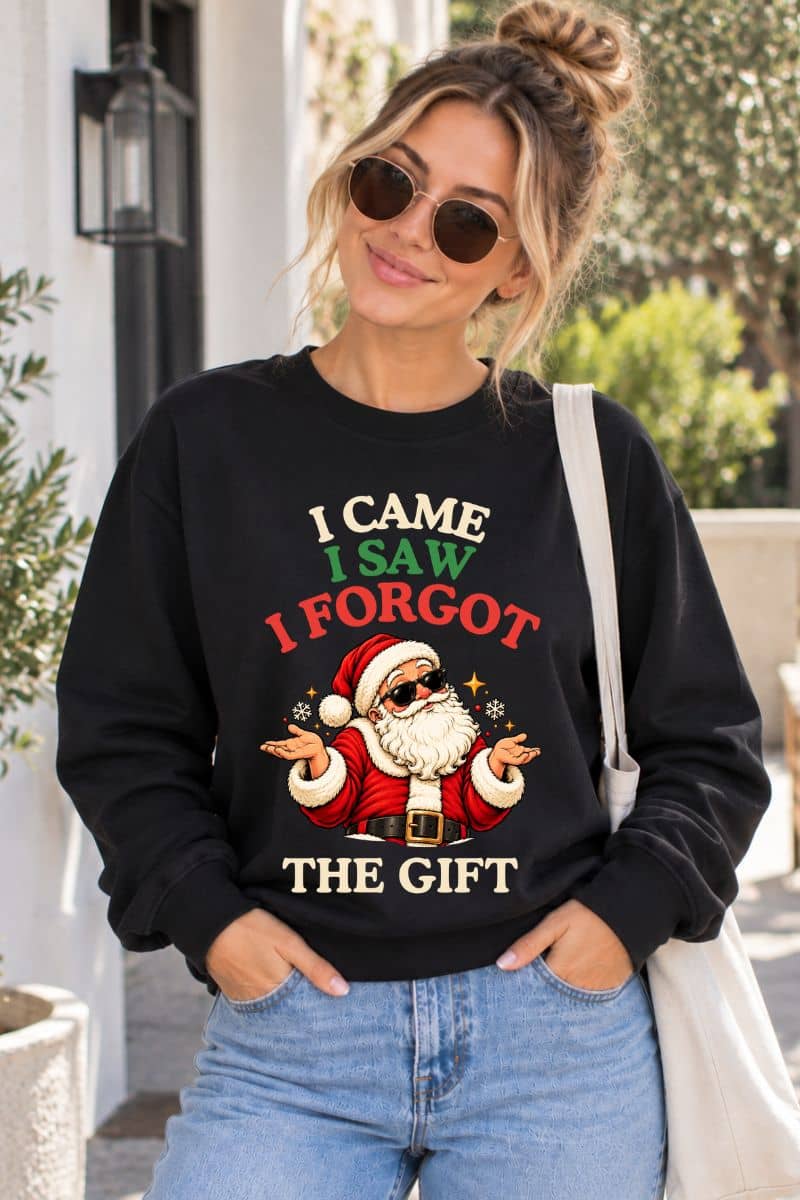Woman in sunglasses wearing a black Christmas Crewneck with a humorous Santa illustration and “I Came. I Saw. I Forgot the Gift” design outdoors.