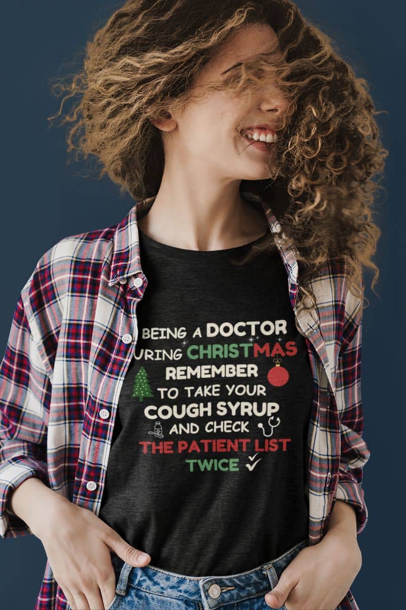 christmas doctor graphic tee with a  person wearing a black t-shirt with text 'Being a doctor during Christmas, remember to take your cough syrup and check the patient list twice', standing against a dark background