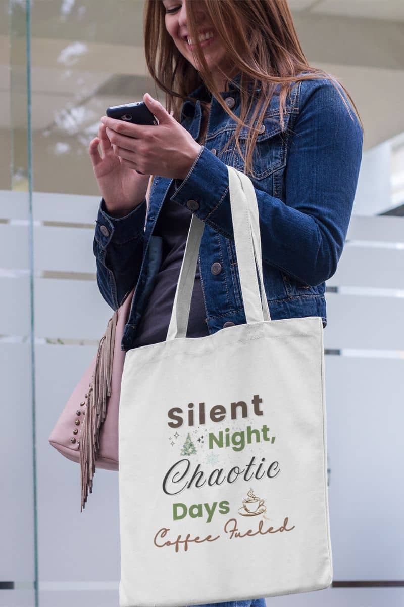 christmas gift bag with a person holding a tote bag with text 'Silent Night, Chaotic Days, Coffee Fueled' and a phone, standing against a glass door.
