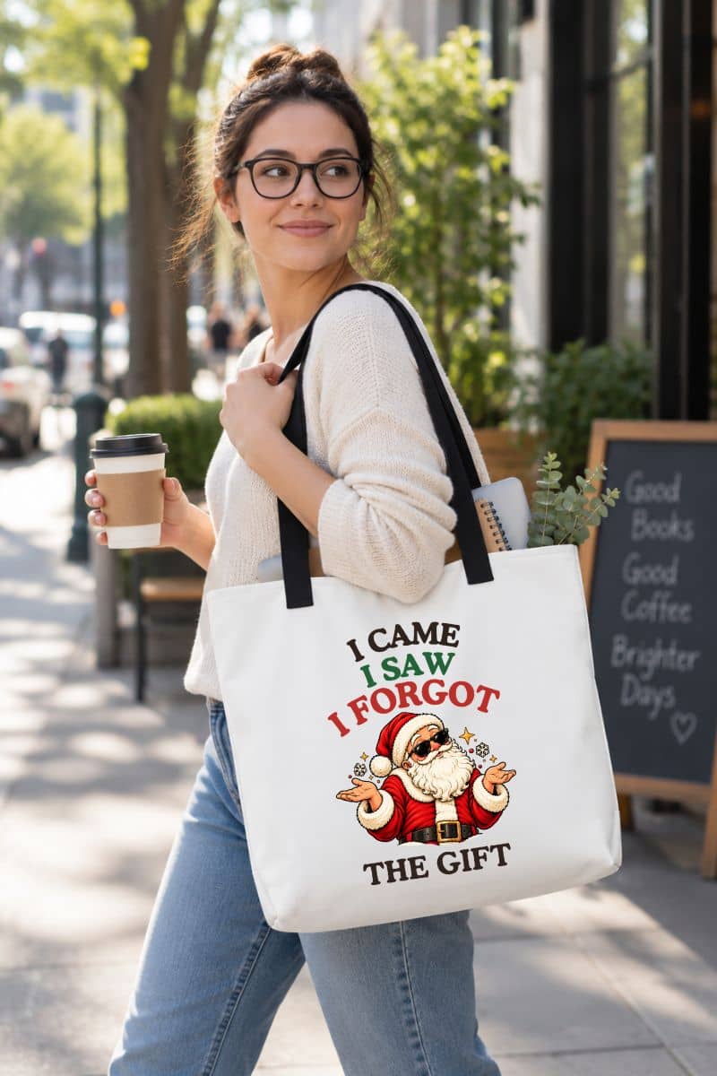 Woman in a cream sweater carrying a Christmas gift bag with black handles, featuring a shrugging Santa Christmas graphic, a notebook, and greenery inside.