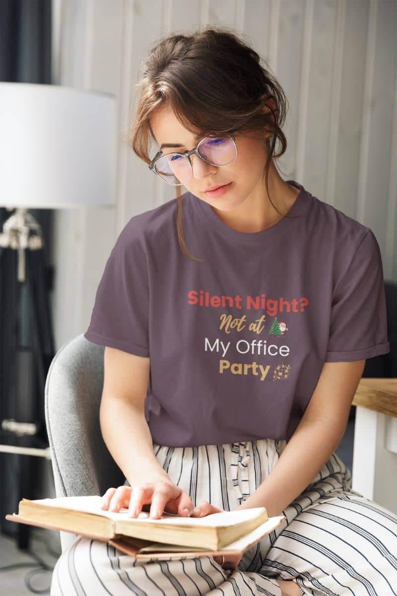 christmas humor shirt with a woman wearing a t-shirt with humorous text 'Silent Night? Not at My Office Party', sitting on a chair reading a book.
