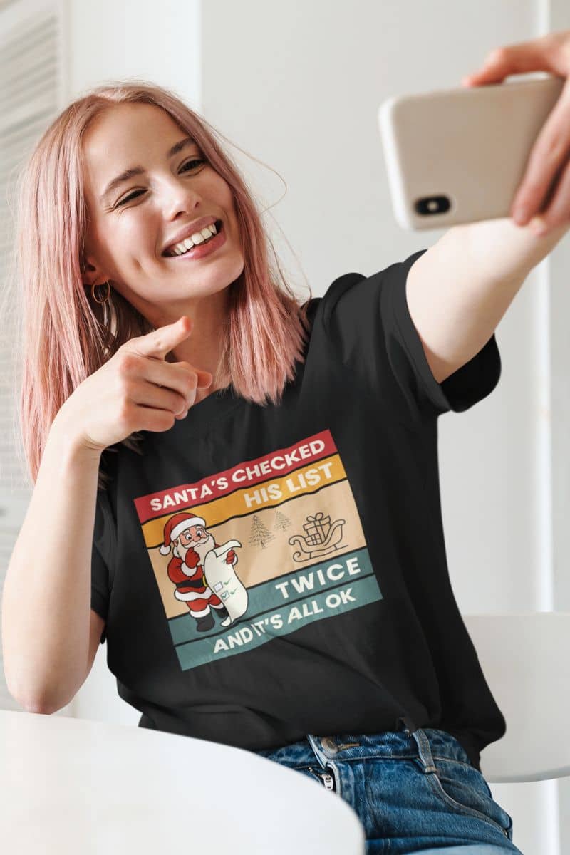 christmas humor shirt with a person wearing a black t-shirt with text 'Santa's Checked His List Twice And It's All Ok', taking a selfie.