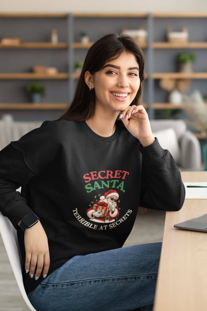 Christmas party sweatshirt featuring a funny Secret Santa design on a black crewneck worn by a woman sitting at a desk.