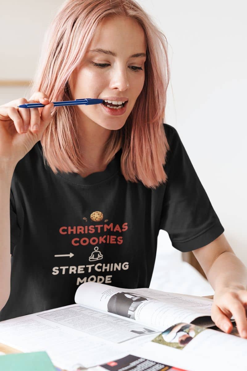 christmas self care shirt with a person wearing a black t-shirt with text 'Christmas Cookies, Stretching Mode', holding a pen and looking at a magazine.