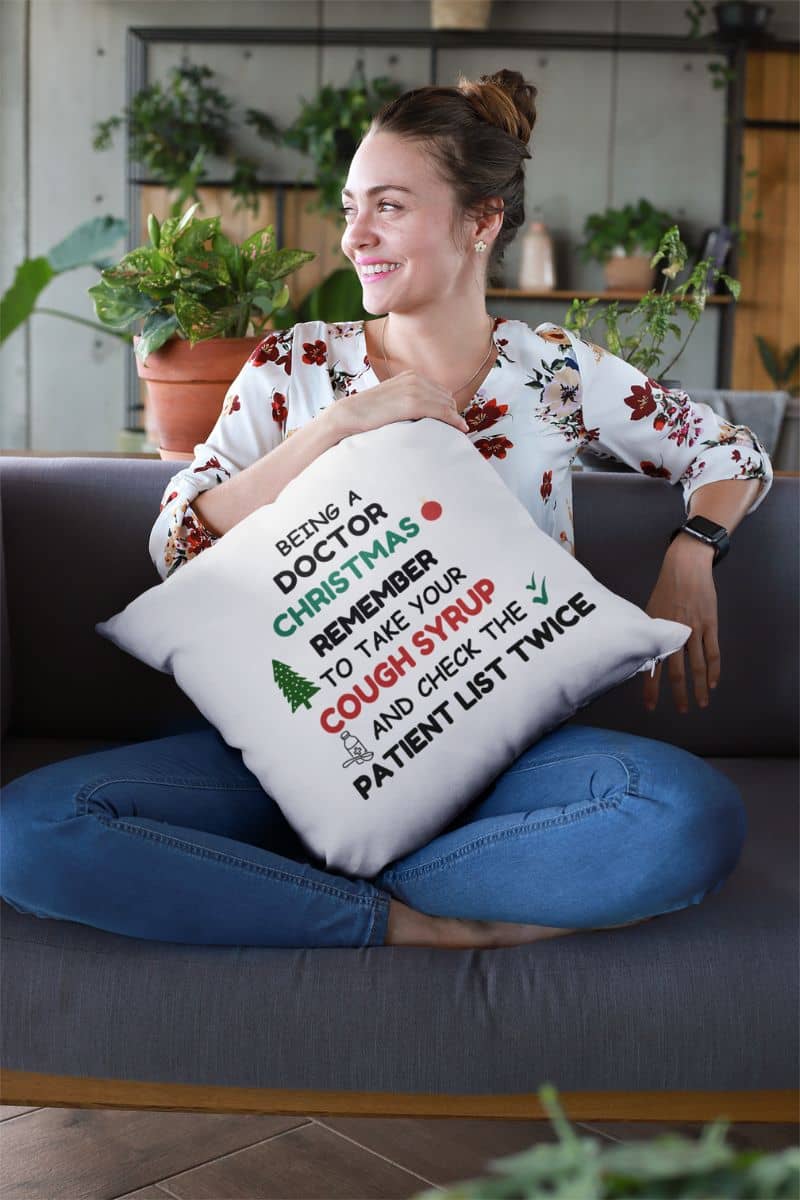 christmas sofa pillow with a woman sitting on a couch holding a pillow featuring the text 'Being a doctor during Christmas, remember to take your cough syrup and check the patient list twice'