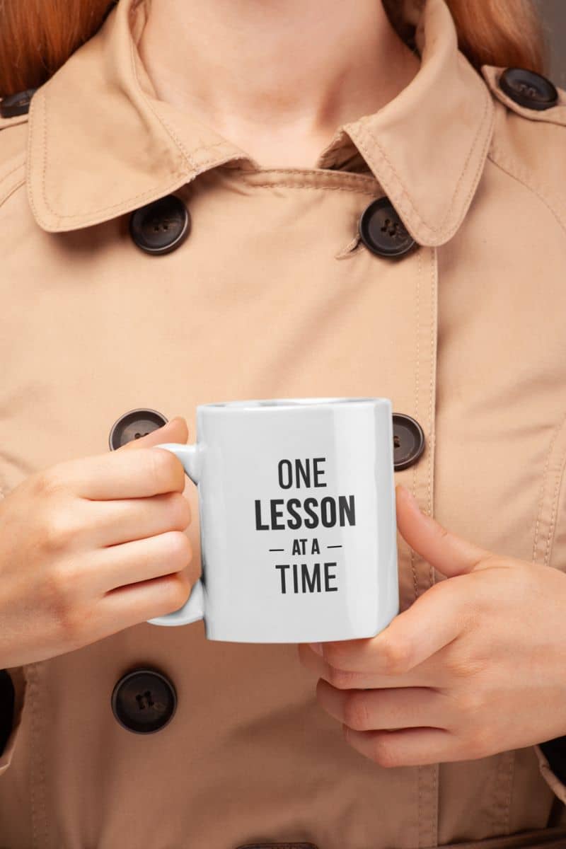 classroom coffee mug with a person in a beige trench coat holds a white ceramic mug with the black printed text 'ONE LESSON AT A TIME'.