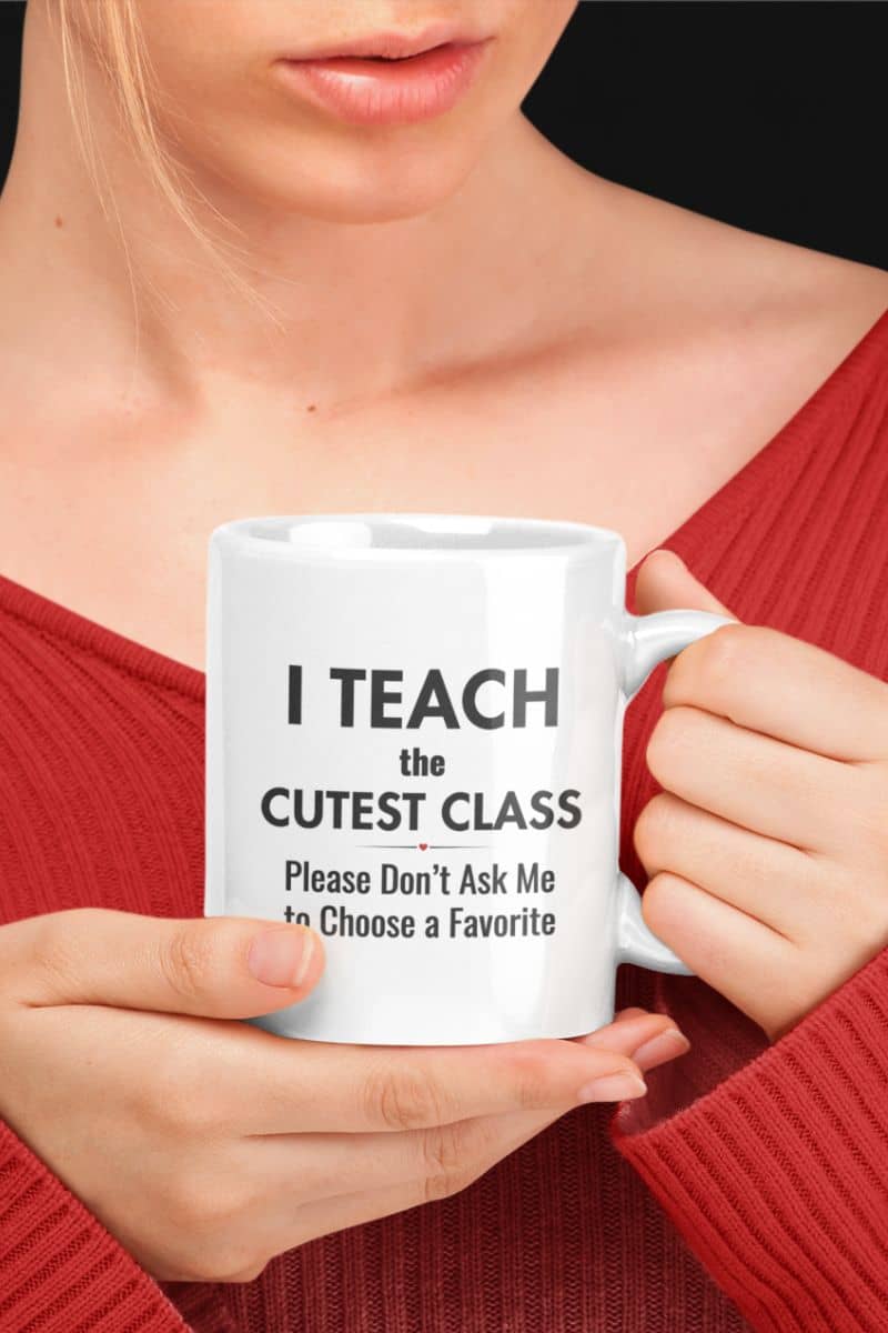 Classroom mug held by a person in a red ribbed sweater, featuring the text “I Teach the Cutest Class. Please Don’t Ask Me to Choose a Favorite” against a dark background.