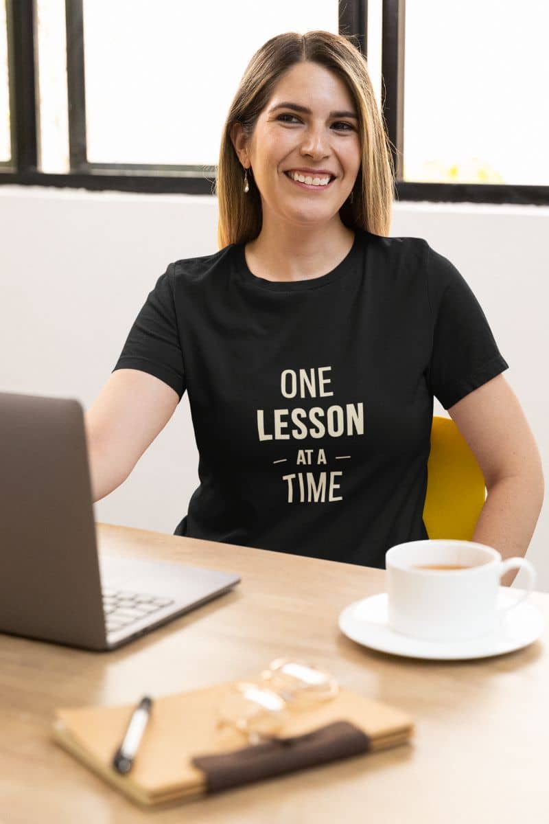 classroom shirt with a woman smiling at a desk, wearing a black t-shirt that reads 'ONE LESSON AT A TIME' 