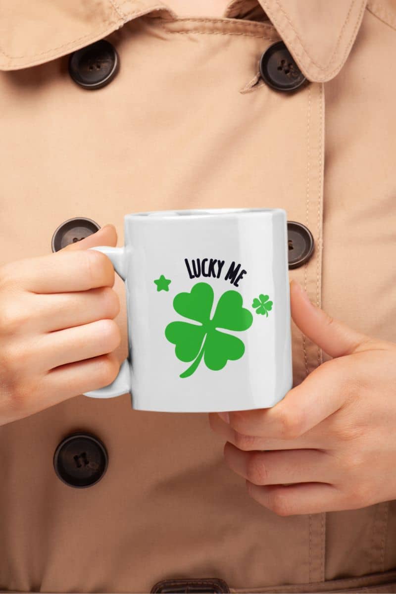 clover coffee mug with a person holding a mug with a four-leaf clover design and 'Lucky Me' text, wearing a beige coat.