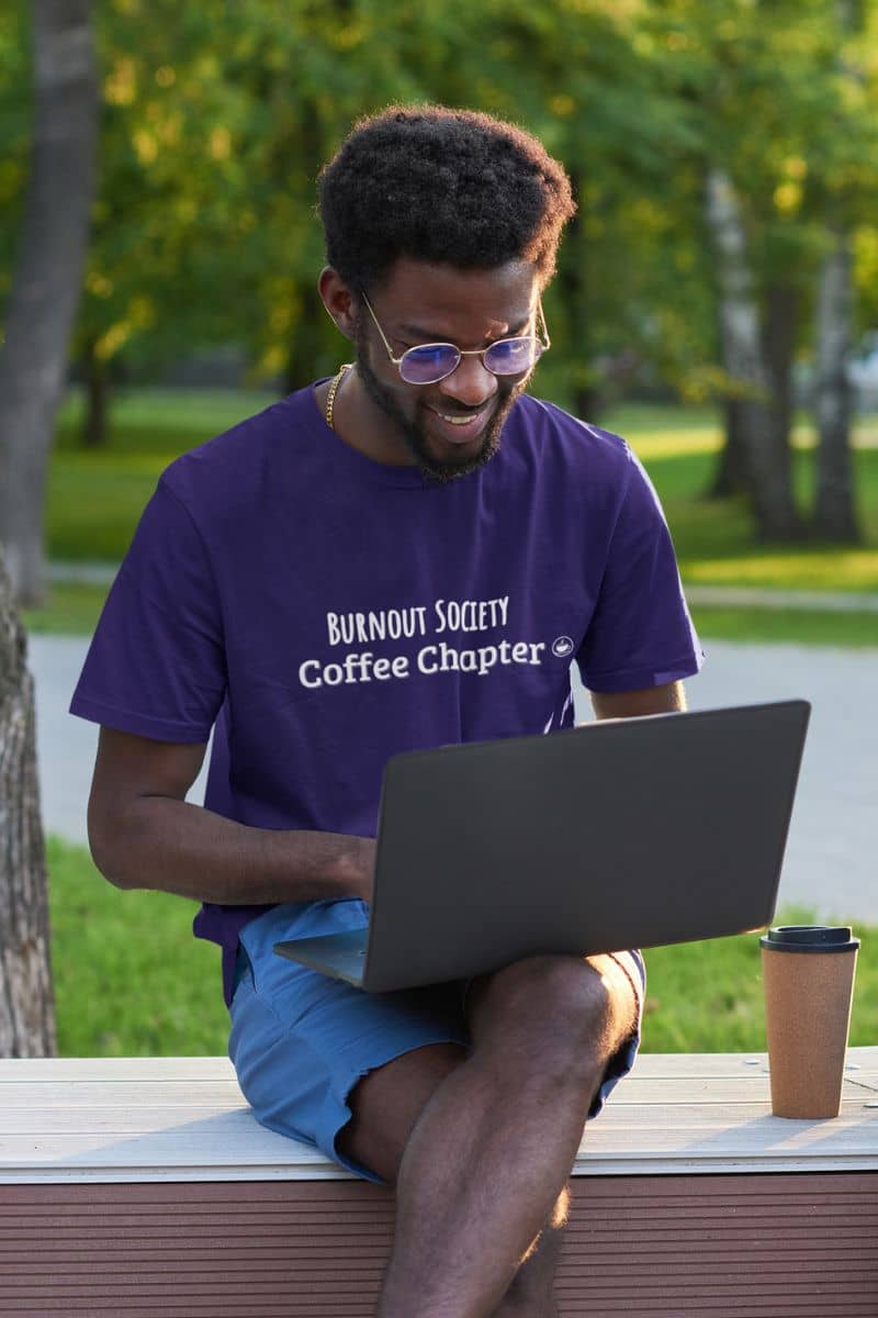 coffee addict work tee with a man in a purple 'Burnout Society Coffee Chapter' t-shirt using a laptop outdoors.