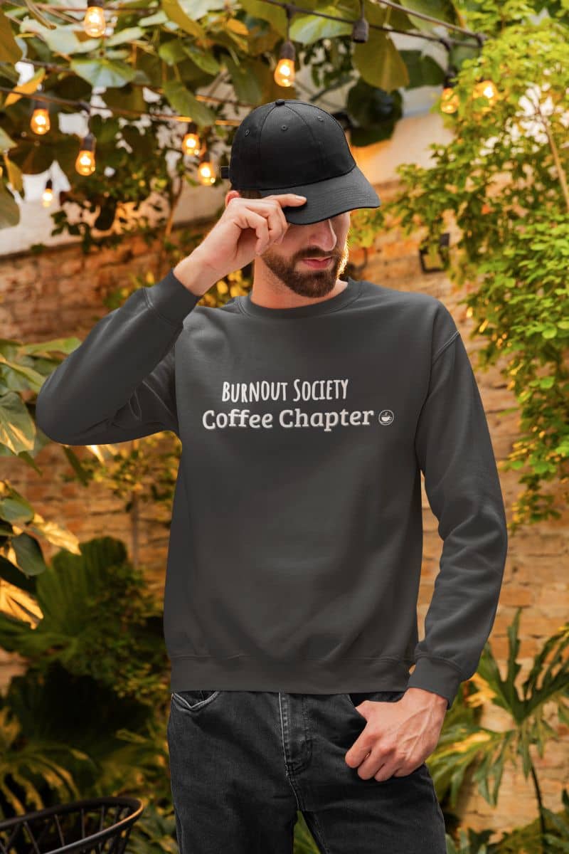 coffee chapter hoodie with a man wearing a dark gray sweatshirt with 'Burnout Society Coffee Chapter' text, standing outdoors with greenery and lights in the background.