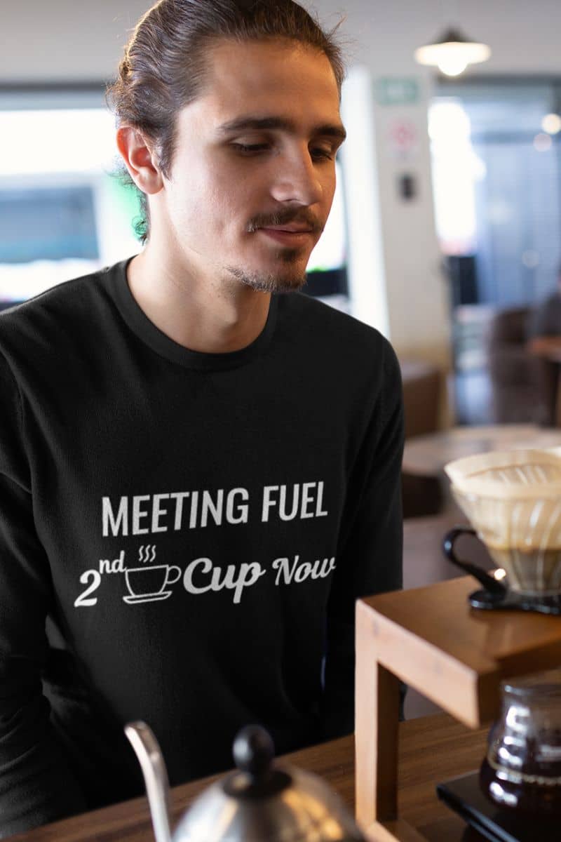 coffee humor sweatshirt with a man wearing a black sweatshirt with text 'Meeting Fuel: 2nd Cup Now' in a coffee shop setting
