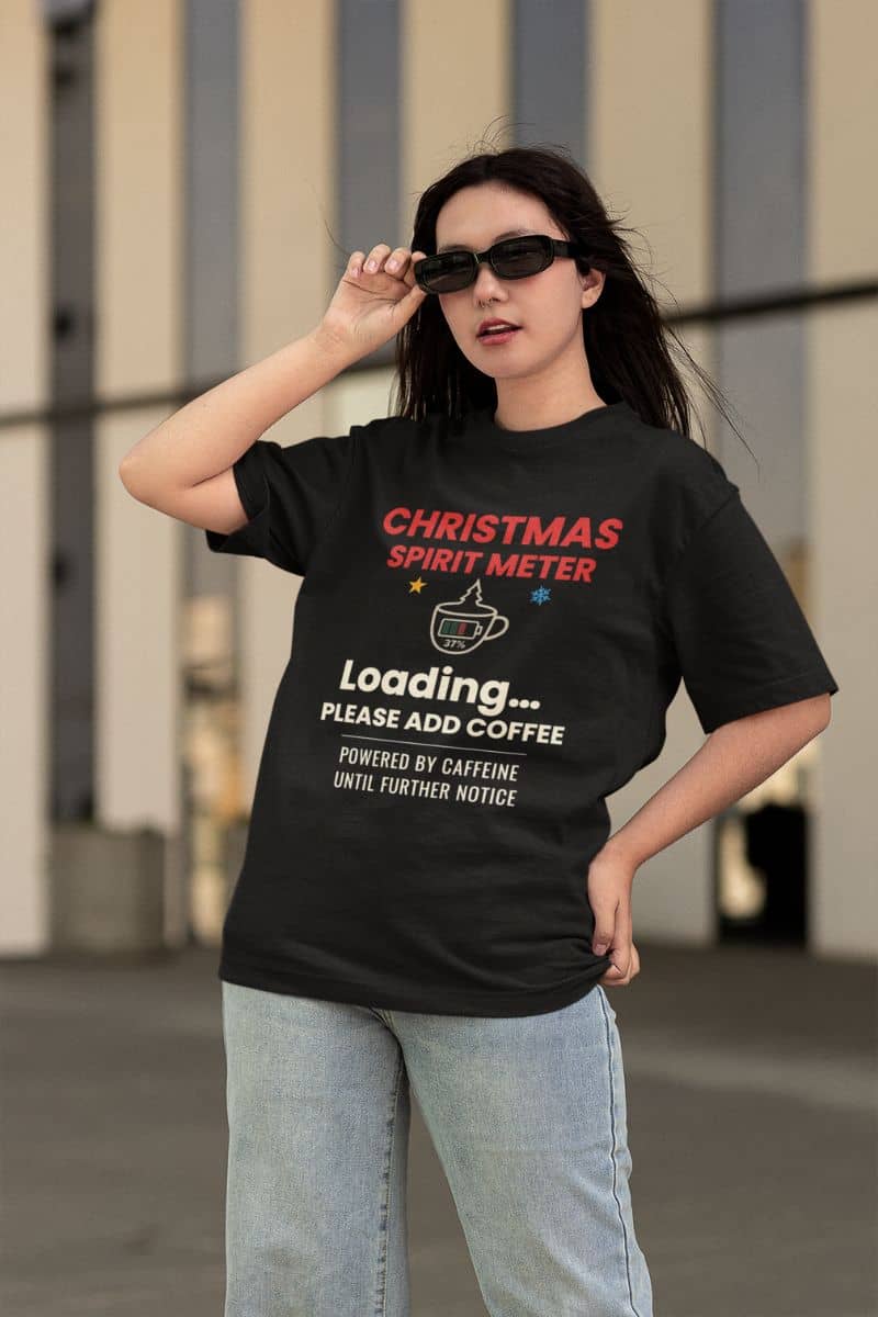 coffee lover christmas shirt with a person wearing a black t-shirt featuring the text 'Christmas Spirit Meter, Loading…37%. Please add coffee. Powered by caffeine until further notice', standing outdoors.
