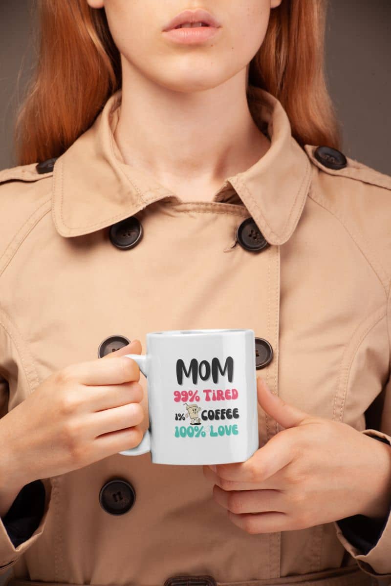 coffee lover mom mug with a person holding a mug with text 'Mom 99% Tired, 1% Coffee, 100% Love' in a beige coat against a neutral background