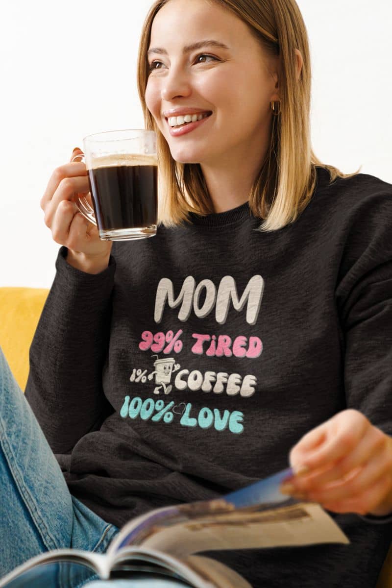 coffee lover mom sweatshirt with a woman wearing a black sweatshirt with text Mom 99% Tired, 1% Coffee, 100% Love', holding a cup of coffee and reading a magazine.