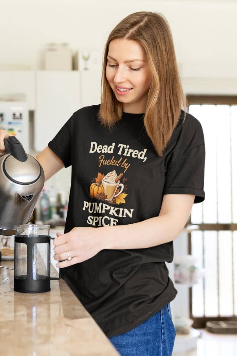 coffee lover shirt with a woman in a kitchen wearing a black graphic t-shirt that reads 'Dead Tired, Fueled by Pumpkin Spice' with an illustration of a pumpkin and a mug. She is pouring hot water from a kettle into a French press.