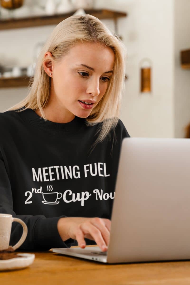 coffee lover sweatshirt with a woman using a laptop with a black sweatshirt labeled 'Meeting Fuel: 2nd Cup Now' in a kitchen setting.