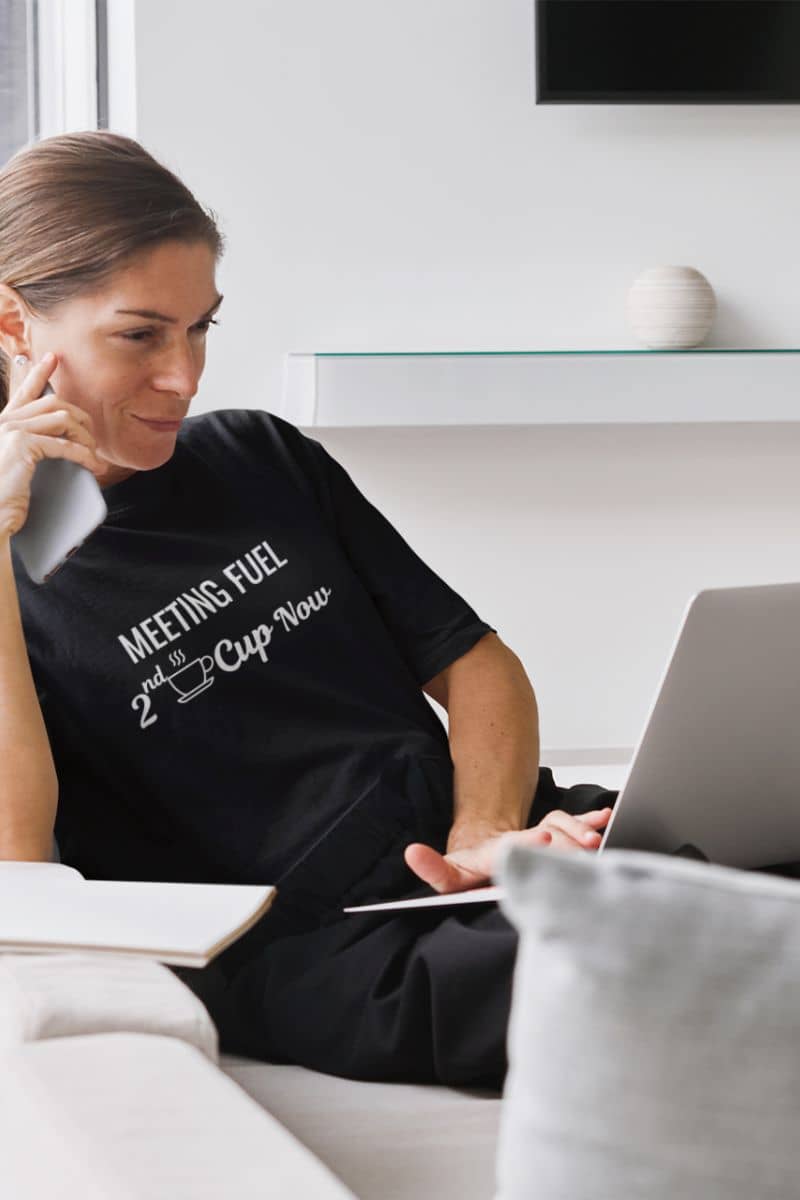 coffee lover tee with a woman sitting on a couch using a laptop, wearing a black t-shirt with text 'Meeting Fuel: 2nd Cup Now'.
