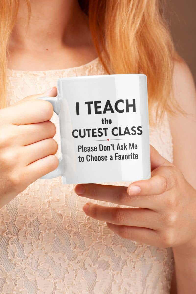 Coffee mug for teachers held by a person, featuring the text “I Teach the Cutest Class. Please Don’t Ask Me to Choose a Favorite” in black lettering.