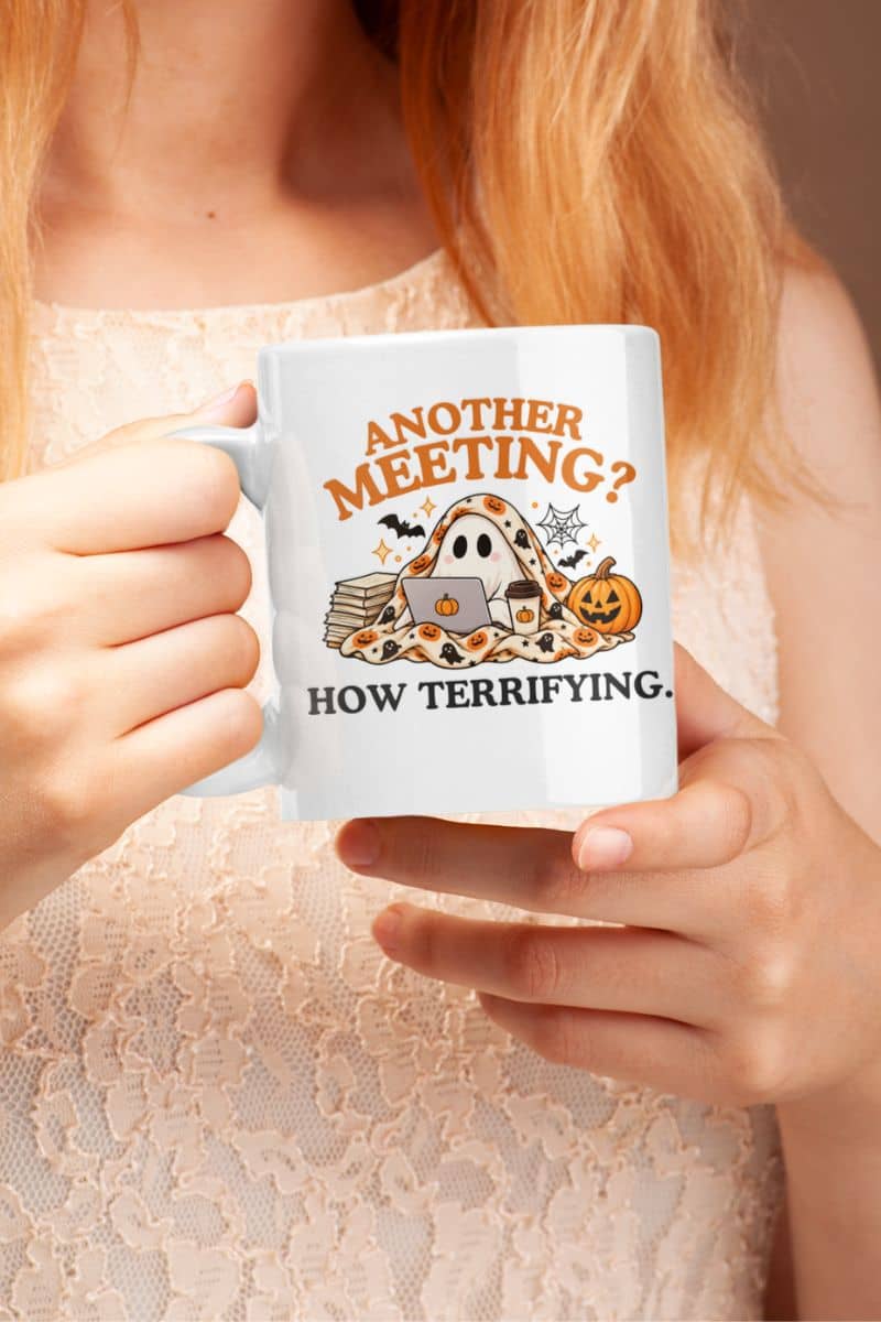 A person in a beige lace top holding a coffee mug for work with a Halloween-themed graphic that reads 'Another Meeting? How Terrifying.'