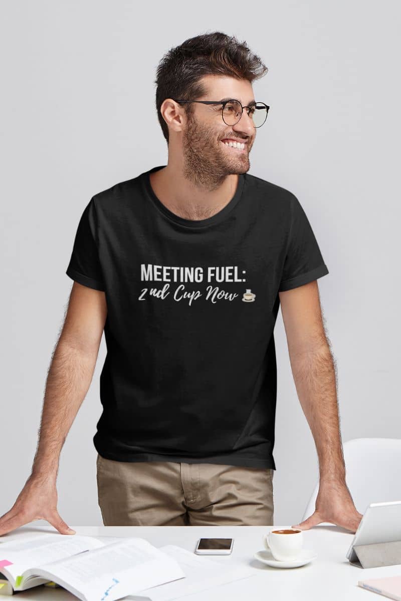 Coffee Quote Tee with a man in a black T-shirt featuring the text 'Meeting Fuel: 2nd Cup Now', standing at a desk.