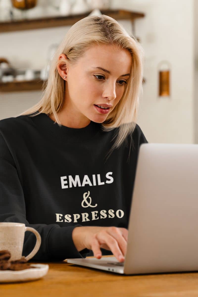 coffee sweatshirt with a woman wearing a black sweatshirt with text 'EMAILS & ESPRESSO', using a laptop in a kitchen.