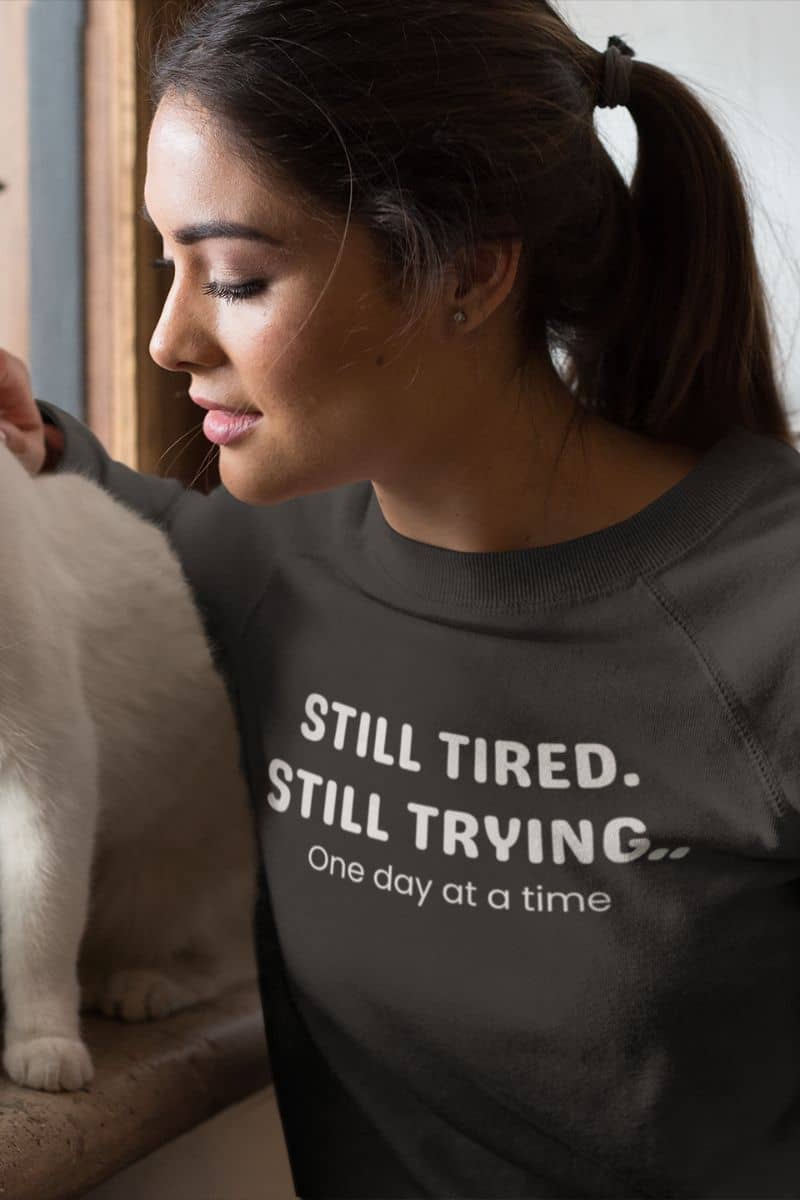 comfort motivation sweatshirt with a  woman wearing a sweatshirt with text 'Still Tired. Still Trying. One day at a time', petting a cat indoors.
