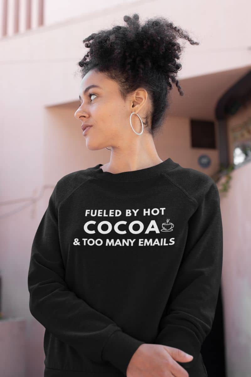 comfy office sweatshirt with a person wearing a black sweatshirt with text 'Fueled by hot cocoa & too many emails', standing outdoors.