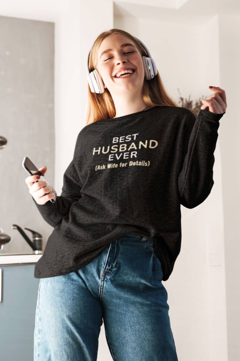couple sweatshirt with a person wearing a black sweatshirt with text 'Best Husband Ever. (Ask Wife for Details)', listening to music in a kitchen.
