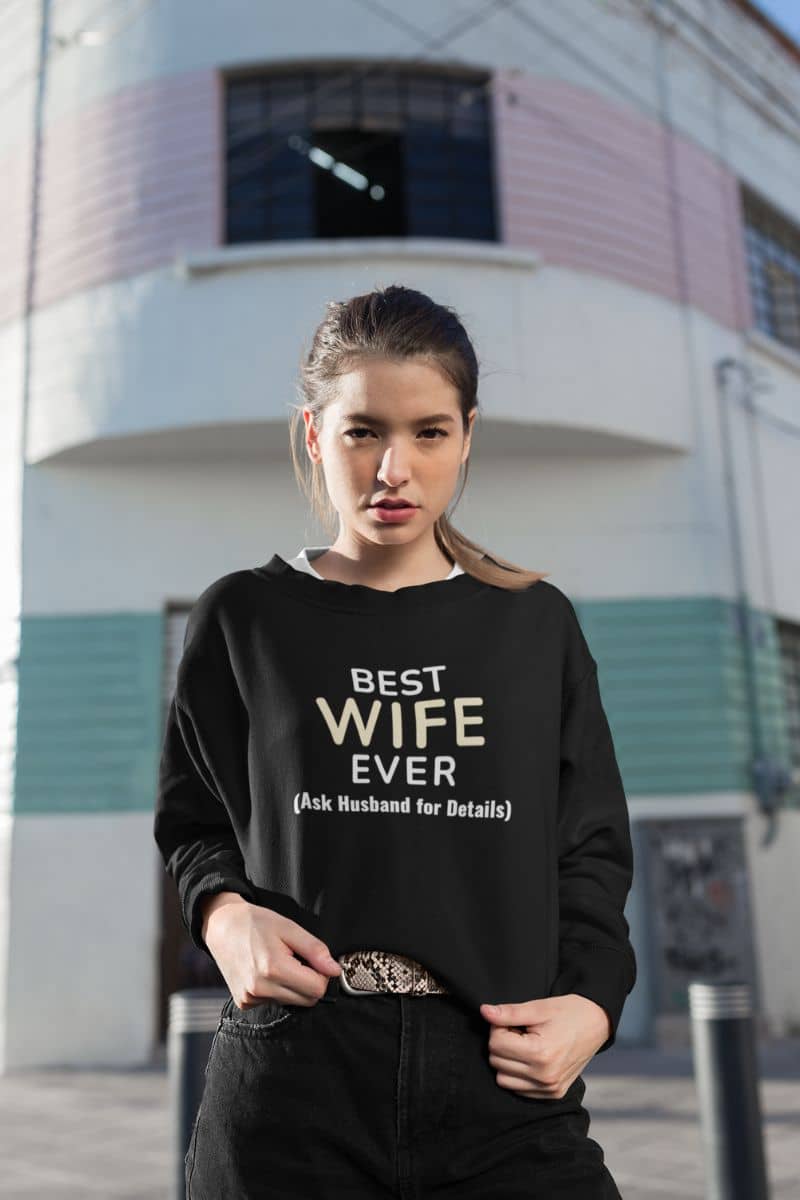 couple sweatshirts with a woman wearing a black sweatshirt with text 'Best Wife Ever. (Ask Husband for Details)', standing in front of a building.