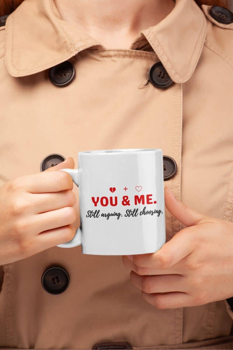 couples daily coffee cup with a person holding a mug with text 'YOU & ME. Still arguing. Still choosing.' against a beige coat background.