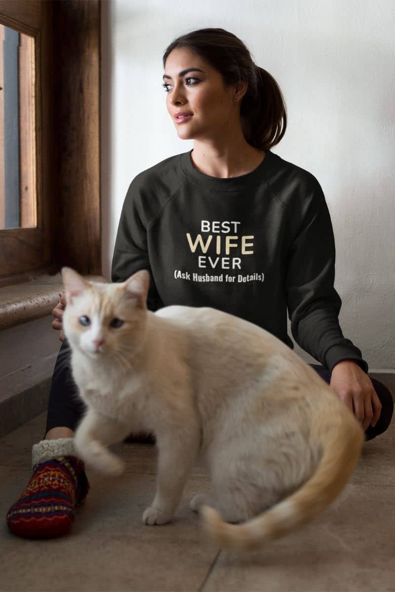 Woman wearing a sweatshirt with text 'Best Wife Ever. (Ask Husband for Details)', sitting with a white cat.